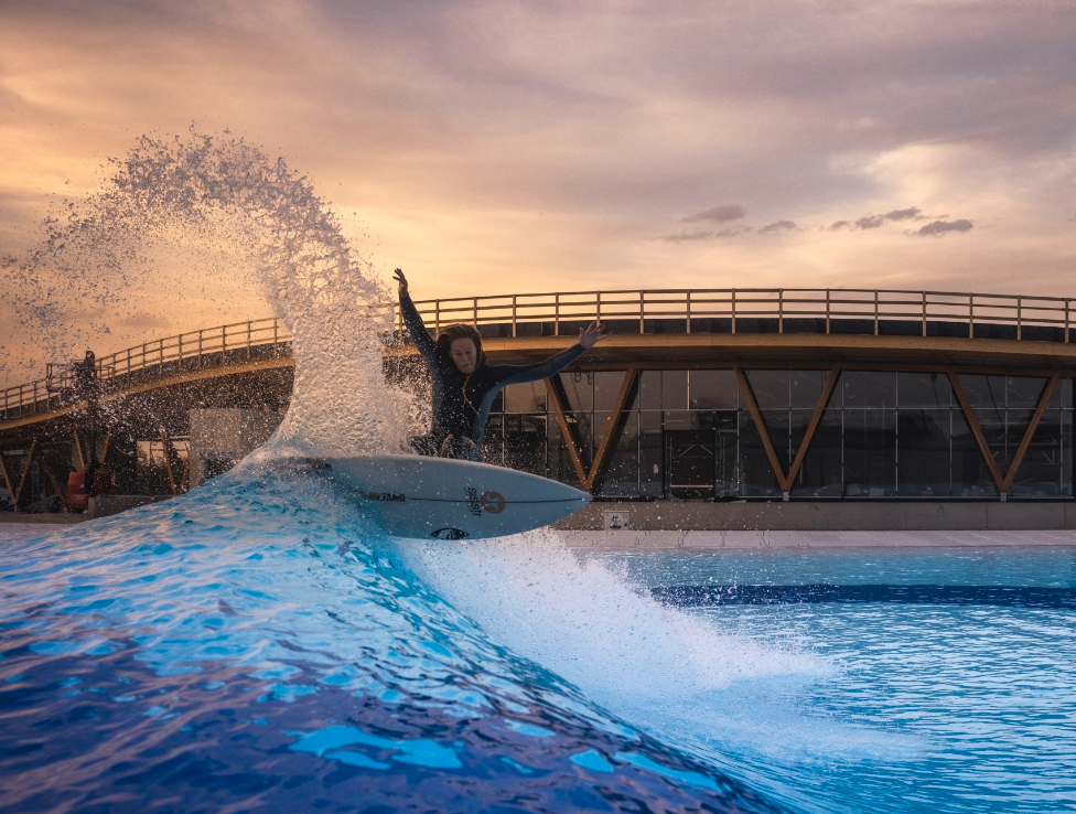 A surfer in a wetsuit rides a wave in an artificial pool, water spraying behind him, with a modern building and the warm glow of sunset framing the scene—capturing the vibrant energy of Santa Clarita Valley.